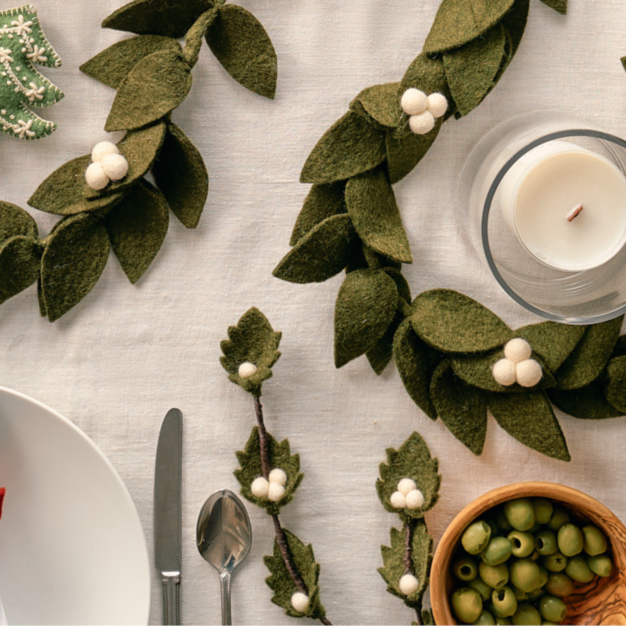 Handmade, felt green leaf garland with white berries on a table setting with cutlery and a candle. Fair trade | Global Goods Partners
