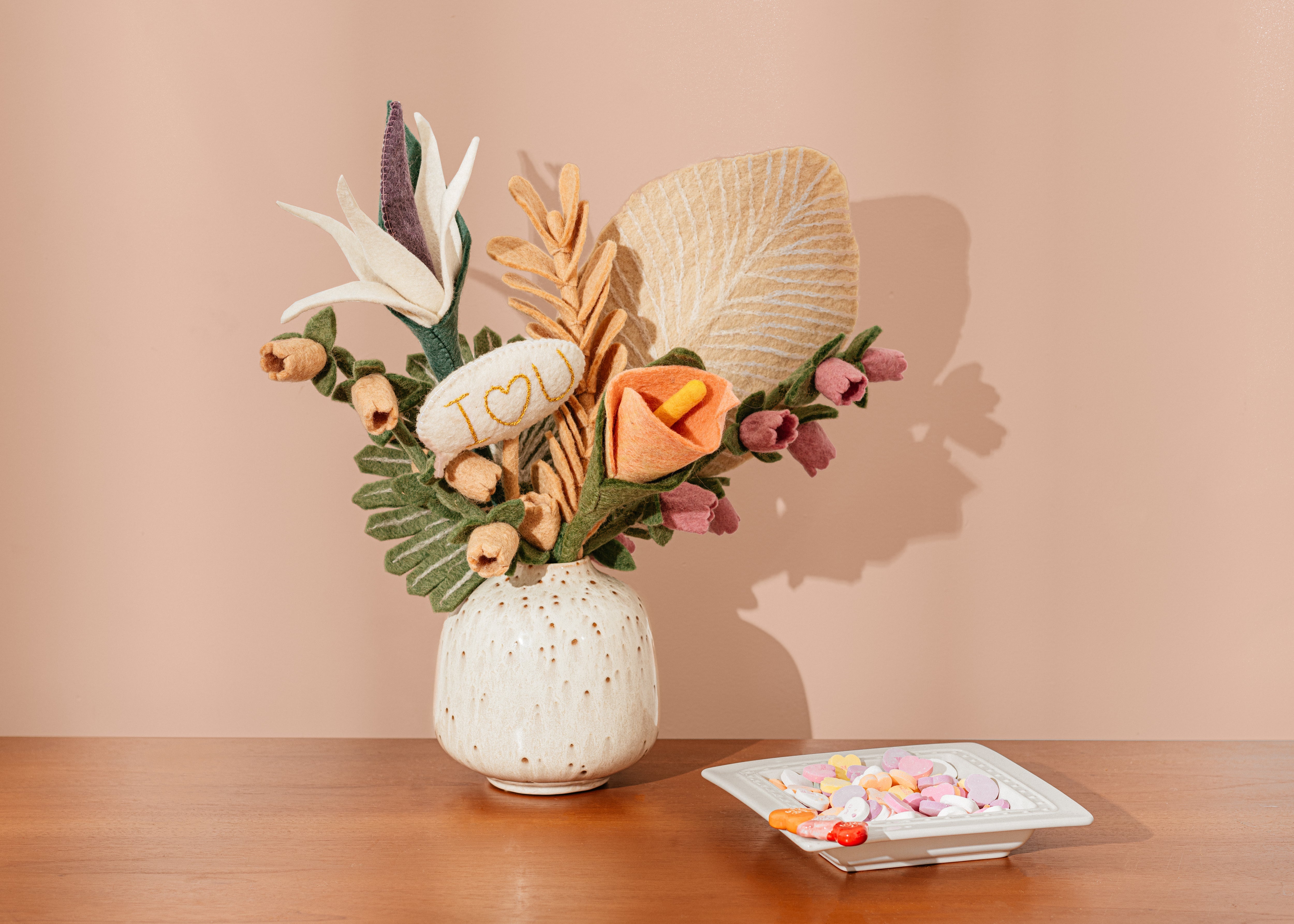 Bouquet of vibrant felt flowers on a table with candy hearts and a pink background