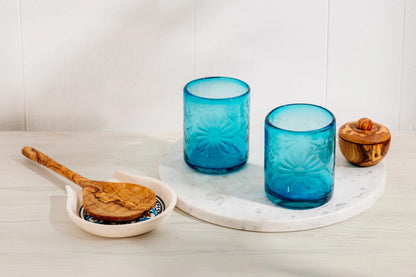 Two blue glass tumblers on a white marble tray with wooden items on a light wooden surface. Fair trade recycled glass | Global Goods Partners