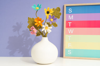 White vase with colorful handful felted flowers against a purple wall with a calendar, all fair trade | Global Goods Partners
