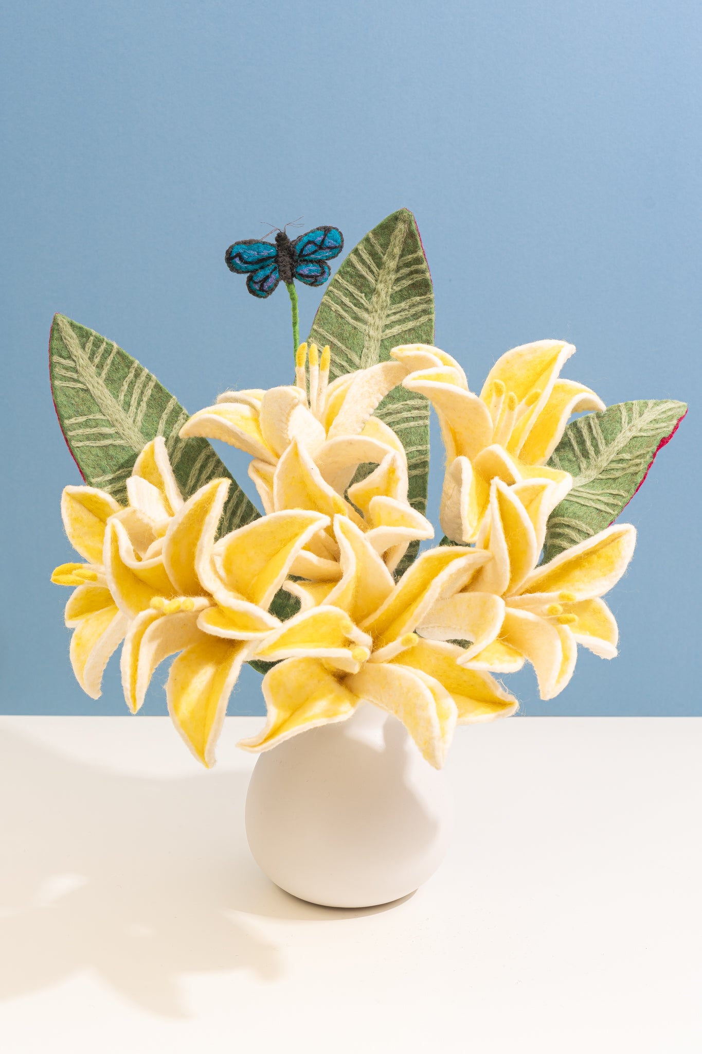 Yellow fair trade felt flowers in a white vase with green leaves and a blue butterfly on a light blue background | Global Goods Partners