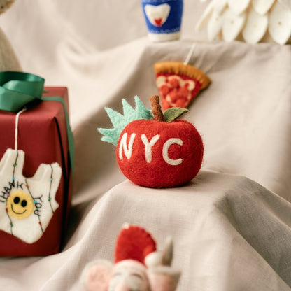 Handmade, fair trade, felt red apple ornament with 'NYC' on a soft surface. 
