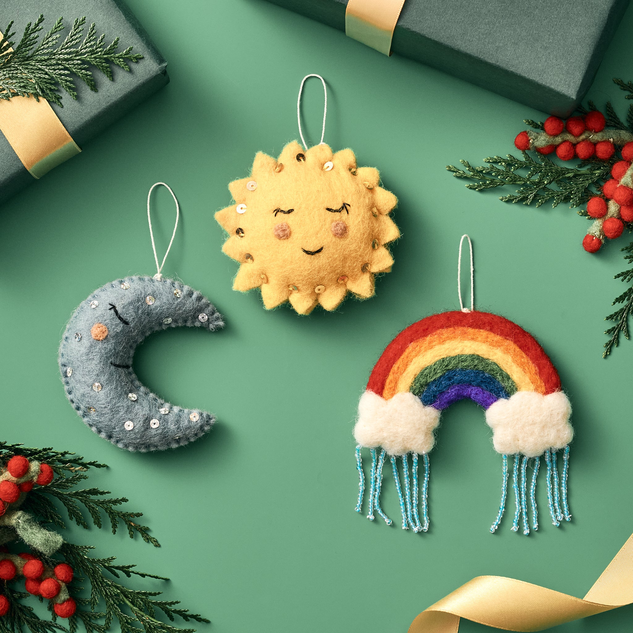 Handmade, fair trade felt ornaments shaped like a sun, moon, and rainbow on a green background with gift boxes and berries | Global Goods Partners