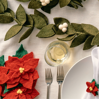 Christmas-themed table setting with handmade felt poinsettia flowers and holly leaves on a white tablecloth | Global Goods Partners