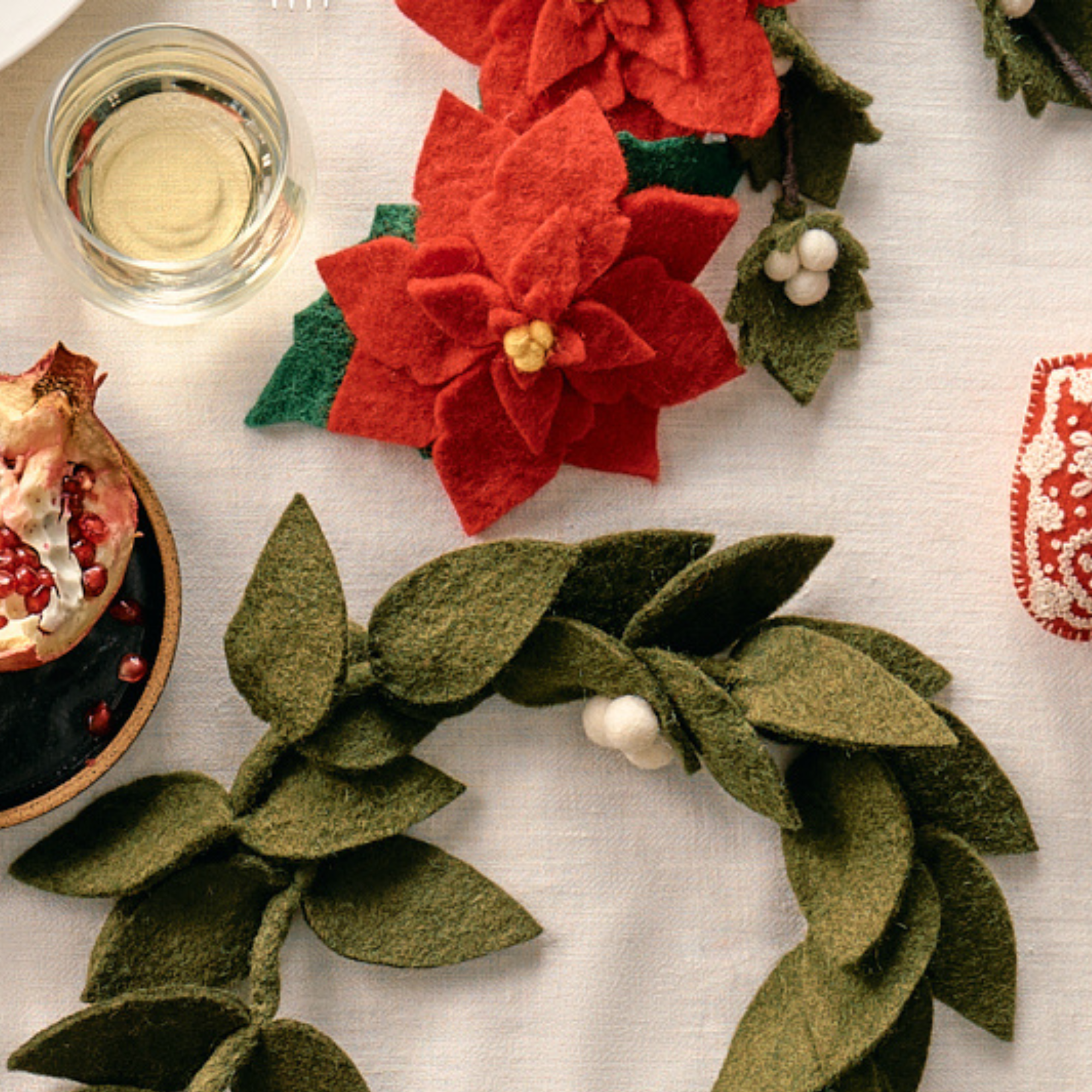 Fair trade felt poinsettia flowers and green leaf wreath on a textured surface with a glass of white wine | Global Goods Partners