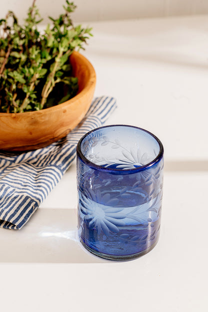 Blue glass tumbler with floral patterns on a white surface with a wooden bowl and striped cloth. Recycled glassware handmade in Mexico | Global Goods Partners