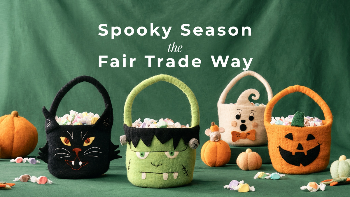 Four handmade Halloween-themed felt candy baskets arranged on a green fabric backdrop with pumpkins and scattered candy. The baskets are black with a cat face, green with a monster face, cream with a ghost face and bow tie, and orange with a pumpkin face. | Global Goods Partners