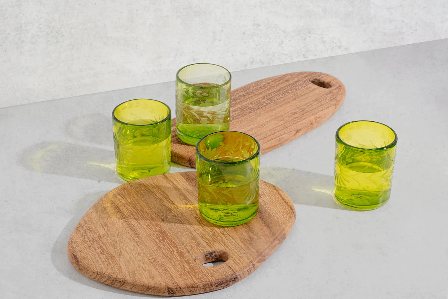 Four green glass tumblers on wooden serving boards against a light background; recycled glass handmade in Mexico | Global Goods Partners