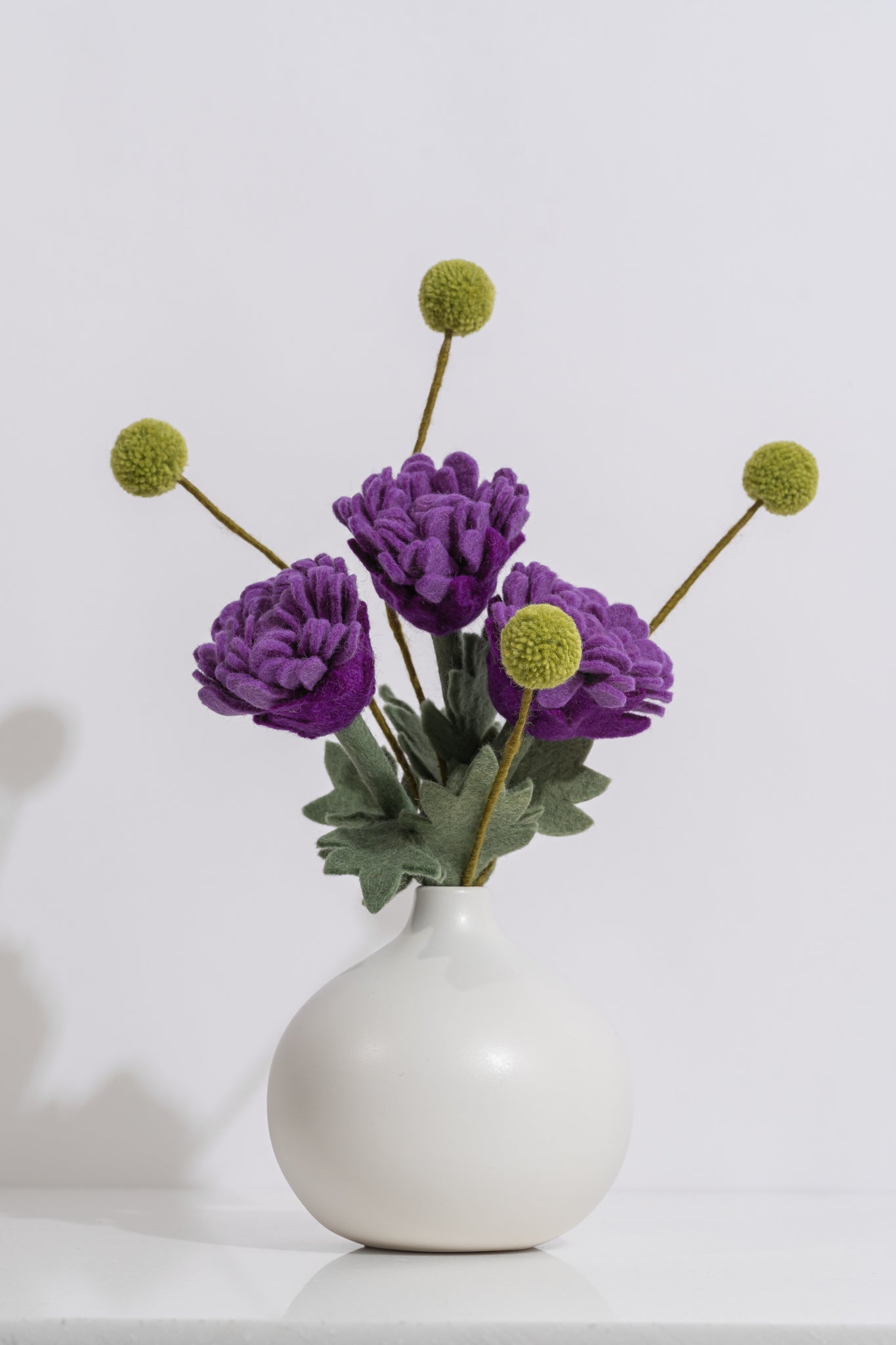 White vase with vibrant purple flowers and thin green stems on a white background. Felt flowers handmade in Nepal | Global Goods Partners
